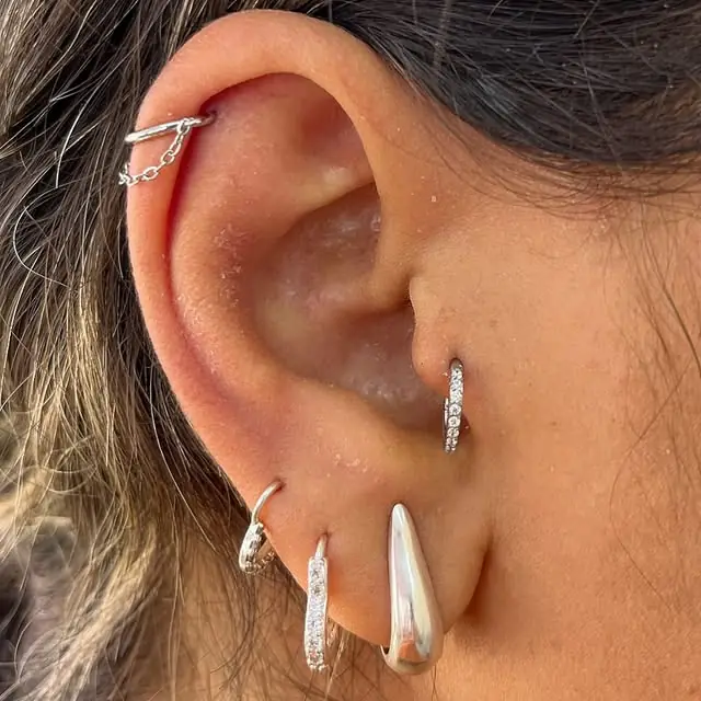 Orelha com piercings prateados: corrente no alto, argola no trago e brincos nos lóbulos